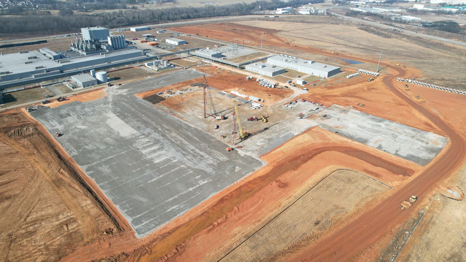 Aerial view of a mission-critical industrial jobsite — mass-graded building pads, deep foundation crane rigs, and stormwater pipe staged alongside an operating plant